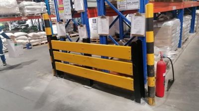Yellow safety barriers installed at the base of racking units in a warehouse, providing protection against impacts, with a fire extinguisher and safety signs visible, provided by Duzi Storage Solutions.